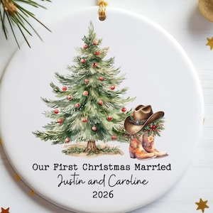 May include: A round white ceramic Christmas ornament featuring a watercolor illustration of a green pine tree decorated with red ornaments, a brown cowboy hat, and a pair of western boots. Text reads: Our First Christmas Married Justin and Caroline 2026.