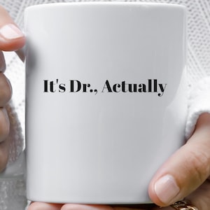 May include: White ceramic mug with the text "It's Dr., Actually" in black. The mug is held by a person wearing gold rings. The background is a white, textured fabric.