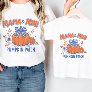 Pode incluir: Duas t-shirts brancas com uma impressão gráfica de uma abóbora laranja com um laço às riscas azuis e brancas, rodeada por flores e folhas. O texto MAMA & MINI PUMPKIN PATCH está escrito em letras cor de laranja e azuis em cada t-shirt.