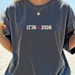 May include: Dark gray t-shirt with the text "1776 2026" in white, with a red star between the numbers. The person is holding a clear plastic cup with iced coffee. The person is wearing a gold necklace and bracelet.