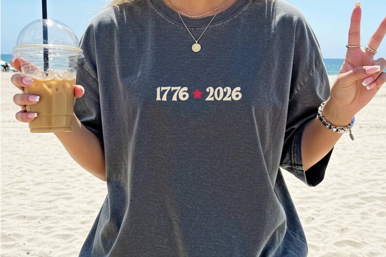 May include: Dark gray t-shirt with the text "1776 2026" in white, with a red star between the numbers. The person is holding a clear plastic cup with iced coffee. The person is wearing a gold necklace and bracelet.