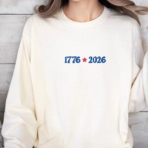 May include: Cream-colored long-sleeve sweatshirt with the text "1776 * 2026" in blue, with a red star. The sweatshirt is made of a soft material and has a crew neck.