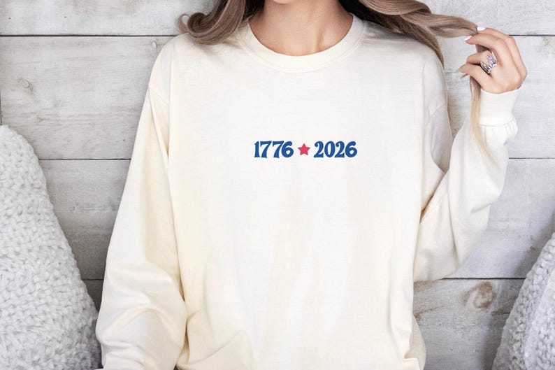 May include: Cream-colored long-sleeve sweatshirt with the text "1776 * 2026" in blue, with a red star. The sweatshirt is made of a soft material and has a crew neck.