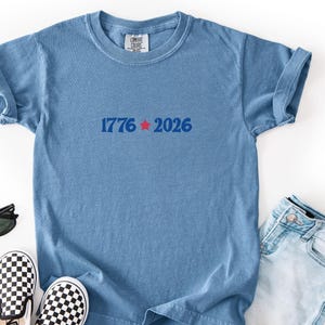 May include: A light blue t-shirt with rolled-up sleeves, featuring the text "1776 2026" in blue with a red star. The shirt is paired with denim jeans, black sunglasses, and checkered slip-on shoes.