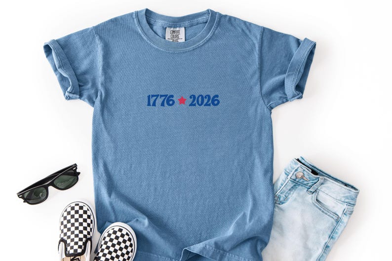 May include: A light blue t-shirt with rolled-up sleeves, featuring the text "1776 2026" in blue with a red star. The shirt is paired with denim jeans, black sunglasses, and checkered slip-on shoes.