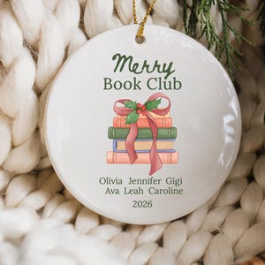 Pode incluir: Um ornamento redondo de cer&acirc;mica branca com uma pilha de quatro livros atados com uma fita vermelha e azevinho. O texto diz Merry Book Club, seguido pelos nomes Olivia, Jennifer, Gigi, Ava, Leah, Caroline e o ano 2026 em verde escuro.