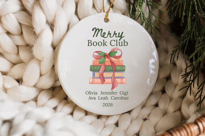 Pode incluir: Um ornamento redondo de cer&acirc;mica branca com uma pilha de quatro livros atados com uma fita vermelha e azevinho. O texto diz Merry Book Club, seguido pelos nomes Olivia, Jennifer, Gigi, Ava, Leah, Caroline e o ano 2026 em verde escuro.