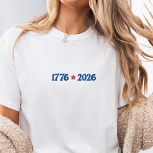 May include: White t-shirt with the text "1776 2026" in blue, with a red star between the numbers. The shirt is a crew neck style. The model is wearing a beige sweater.