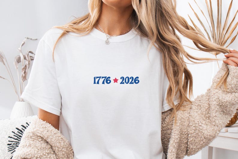 May include: White t-shirt with the text "1776 2026" in blue, with a red star between the numbers. The shirt is a crew neck style. The model is wearing a beige sweater.