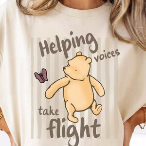 May include: A cream-coloured t-shirt featuring a graphic of a yellow bear and a purple butterfly. The text reads Helping voices take flight in dark brown lettering. The design includes vertical light brown stripes behind the bear and butterfly.