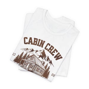 Puede incluir: Dos camisetas blancas dobladas y apiladas. La camiseta superior presenta un gr&aacute;fico marr&oacute;n con las palabras "CABIN CREW" sobre una ilustraci&oacute;n de caba&ntilde;a, monta&ntilde;as y &aacute;rboles. Los n&uacute;meros "20" y "26" est&aacute;n a ambos lados del gr&aacute;fico de la caba&ntilde;a.