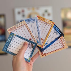 Könnte beinhalten: Eine Hand hält einen Fächer aus Lorepad-Notizblöcken. Jeder Notizblock hat ein anderes Design, darunter Blumenmuster und Zeichentrickfiguren. Die Notizblöcke sind blau, gelb und orange mit dem Text "LOREPAD" oben.