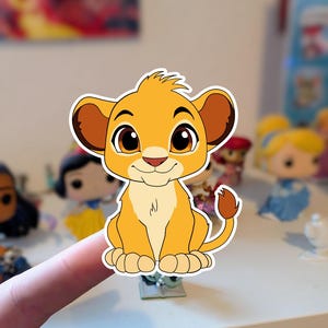 Könnte beinhalten: Ein Aufkleber von Simba aus Der König der Löwen. Der Aufkleber zeigt ein Cartoon-Löwenjunges mit orangefarbenem Fell, braunen Ohren und großen, ausdrucksstarken Augen. Das Junge sitzt mit freundlichem Gesichtsausdruck. Andere Figuren sind im Hintergrund.