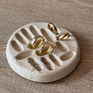 Könnte beinhalten: Ein runder, cremefarbener und beigefarbener Ringhalter mit mehreren goldenen Ringen. Der Ringhalter hat mehrere gebogene Schlitze zur Aufnahme von Ringen. Die Ringe sind in verschiedenen Stilen und Größen, darunter ein Diamantring.