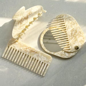 Può includere: Tre accessori per capelli color avorio con un motivo marmorizzato. Il set include un pettine, una forcina e uno specchietto compatto con un pettine su un lato. La parola "Raya" è scritta in oro su ogni articolo.