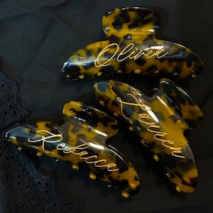 May include: Three tortoiseshell hair clips with gold lettering. The names Olivia, Lauren, and Rebecca are inscribed on the clips.