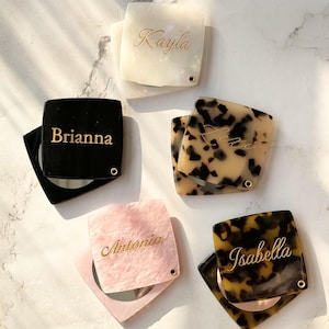 Könnte beinhalten: Eine Sammlung personalisierter Kompaktspiegel in verschiedenen Farben und Mustern. Die quadratischen Spiegel tragen Namen wie "Kayla", "Brianna", "Antonia" und "Isabella" in goldener Schrift. Die Kompakte haben einen kleinen Spiegel im Inneren und eine dekorative Außenschale.