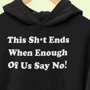 Puede incluir: Una sudadera con capucha negra con texto blanco impreso en el pecho que dice: This Sh*t Ends When Enough Of Us Say No! La prenda incluye una capucha con cordones y un bolsillo tipo canguro, colgada en una percha de madera.