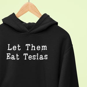 以下が含まれることがあります： 黒いフード付きスウェットシャツに「Let Them Eat Teslas」という白い文字がプリントされています。