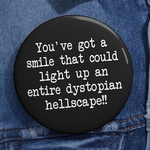 Può includere: Bottone nero con testo bianco che dice "You've got a smile that could light up an entire dystopian hellscape!"