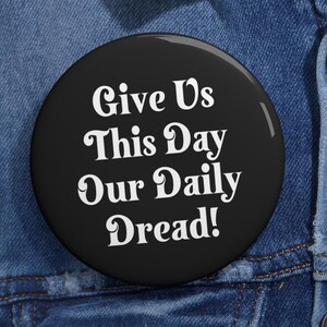 May include: A black button with white text that reads "Give Us This Day Our Daily Dread!"