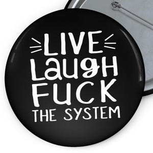 以下が含まれることがあります： 「LIVE LAUGH FUCK THE SYSTEM」と白い文字が書かれた黒いボタン。ボタンの裏にはピンがあり、光沢のある仕上げです。テキストは太字のサンセリフ体です。