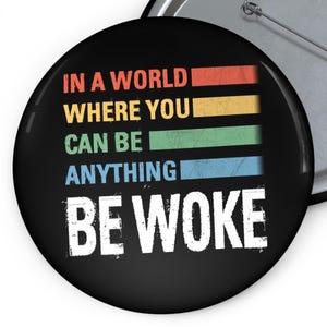 May include: A black button with the text "IN A WORLD WHERE YOU CAN BE ANYTHING BE WOKE" in white. The text is layered over horizontal stripes of red, yellow, green, and blue.