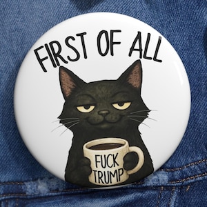 Puede incluir: Un botón blanco con una ilustración de un gato negro sosteniendo una taza de café. La taza tiene el texto "FUCK TRUMP" impreso. Encima del gato está el texto "FIRST OF ALL".