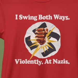 May include: Red t-shirt with the text "I Swing Both Ways. Violently. At Nazis." The design features a clenched fist punching through a stylized swastika, with yellow lightning bolts radiating outwards. The text is in white.