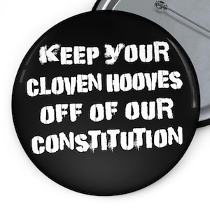 May include: A black button with white text that reads "KEEP YOUR CLOVEN HOOVES OFF OF OUR CONSTITUTION." The button has a pin on the back and a shiny metal edge.