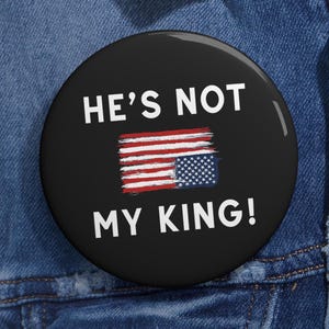 May include: A black button with white text that reads "He's not my king!" and a stylized American flag with the stars and stripes reversed.