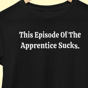 May include: Black t-shirt with the white text "This Episode Of The Apprentice Sucks." The text is centered on the front of the shirt. The shirt is hanging on a wooden hanger.