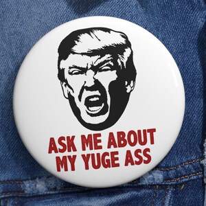 May include: A white button with a black and white image of a man with a serious expression. The text "ASK ME ABOUT MY YUGE ASS" is printed in red below the image.