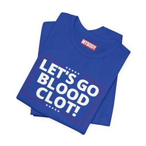Op de afbeelding: Koningsblauw T-shirt met de witte tekst "LET'S GO BLOOD CLOT!" in een rode rechthoek. Het shirt heeft ook de merknaam "MYBODY" in het rood. Het shirt is opgevouwen.