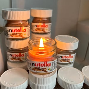 May include: A collection of small glass jars with white lids, labeled "nutella" in red and white. One jar contains a lit orange candle. The jars are arranged in a box, suggesting a gift set or display.