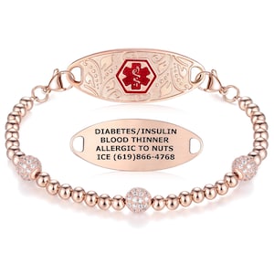 Puede incluir: Pulsera de identificación médica en oro rosa con una cadena de cuentas y dos esferas decorativas con incrustaciones de cristal. La etiqueta ovalada tiene el texto: DIABETES/INSULINA, ANTICOAGULANTE, ALÉRGICO A LOS FRUTOS SECOS, ICE (619)866-4768. La etiqueta principal presenta un símbolo médico rojo.