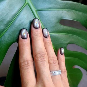 May include: A hand with manicured nails featuring a silver and black design with gold star accents. The nails are square-shaped and have a glossy finish. A silver ring with clear stones is on the ring finger. The background is a large green leaf.