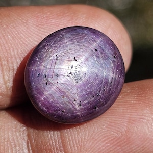 May include: A polished, oval-shaped, purple gemstone with a six-rayed star pattern. The gem has a smooth surface with subtle variations in color and texture. The star effect is created by light reflecting off inclusions within the stone.