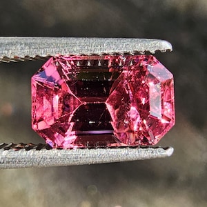 Puede incluir: Una gema rosa rectangular facetada, sujeta por unas pinzas metálicas. La gema tiene múltiples facetas que reflejan la luz, creando un efecto brillante. El color es un rosa vibrante y saturado.