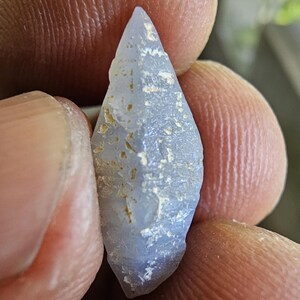 Puede incluir: Una gema en bruto de color azul claro con una punta y una superficie rugosa y texturizada. La piedra tiene pequeñas inclusiones de color marrón dorado y se sostiene entre los dedos. La gema mide aproximadamente 2,5 cm de largo.
