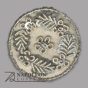 Puede incluir: Un parche bordado circular con una base de tela color crema. Presenta diseños florales y de hojas plateados, con lentejuelas y detalles de cuentas. El borde está adornado con hilo metálico plateado. El logotipo de Napoleon Costume es visible en la parte inferior.