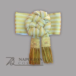 May include: A gold and white decorative sash with a woven knot in the center and two decorative tassels. The sash has a striped pattern and is trimmed with gold detailing. The tassels are gold and have decorative gold caps.
