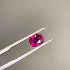 May include: A vibrant pink gemstone, likely a ruby, is held in tweezers. The gemstone is round and has a smooth, polished surface.
