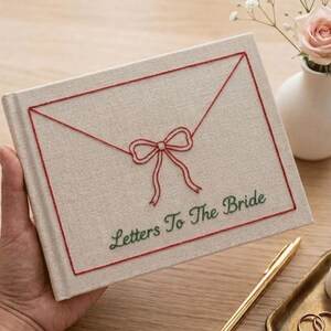May include: A rectangular, light beige fabric-covered book featuring red embroidered lines forming an envelope shape with a central bow. The text Letters To The Bride is embroidered in green thread below the envelope design on the front cover.
