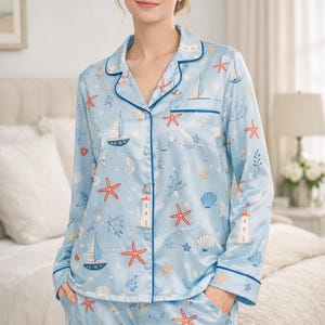 May include: A light blue long-sleeve pajama set featuring a nautical print of sailboats, red starfish, white lighthouses, and seashells. The button-down top has a notched collar, a single chest pocket, and dark blue piping along the edges.