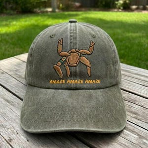 May include: An olive green baseball cap with a distressed fabric finish. The front features an embroidered tan crab-like creature with a small green accent on one leg. Below the embroidery, the text AMAZE AMAZE AMAZE is stitched in yellow.