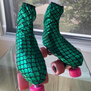Op de afbeelding: Een paar rolschaatsen met groen en zwart geschubde hoezen. De rolschaatsen hebben roze wielen en teenstops. De hoezen zijn gemaakt van een glanzende, getextureerde stof. De rolschaatsen staan voor een raam.