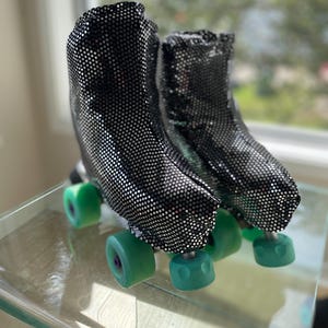 Op de afbeelding: Een paar rolschaatsen met zwarte laarzen met pailletten en groene wielen. De rolschaatsen staan op een glazen oppervlak, met een vage achtergrond van groen. De laarzen zijn bedekt met kleine, reflecterende pailletten.