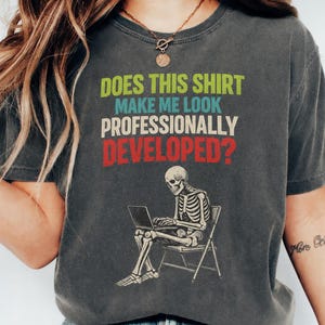 Peut inclure: Un t-shirt gris anthracite à manches courtes avec un motif représentant un squelette assis sur une chaise pliante devant un ordinateur portable. Le texte indique : DOES THIS SHIRT MAKE ME LOOK PROFESSIONALLY DEVELOPED ? en vert, bleu, blanc et rouge.