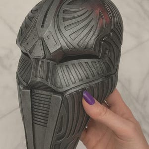 May include: A dark grey, metallic-finished full-face mask featuring intricate, raised geometric patterns and recessed lines across the entire surface. The mask has horizontal eye slits and a vertical, segmented design on the lower face area.